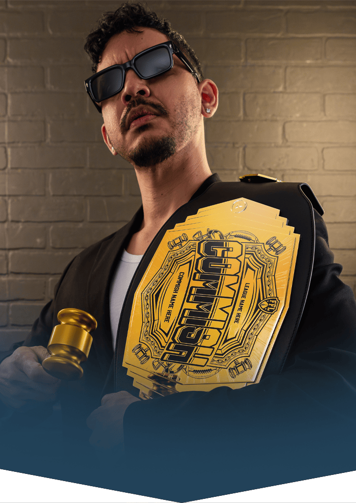 A man wearing black sunglasses, a black blazer, and a white t-shirt holds a gold championship belt with a gavel in his hand, set against a brick wall background.