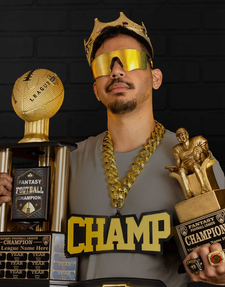 A man wearing a gold crown, gold shield glasses, and a chunky gold chain holds two gold championship trophies against a dark brick wall. He is wearing a gray t-shirt with 'CHAMP' printed in bold gold letters.